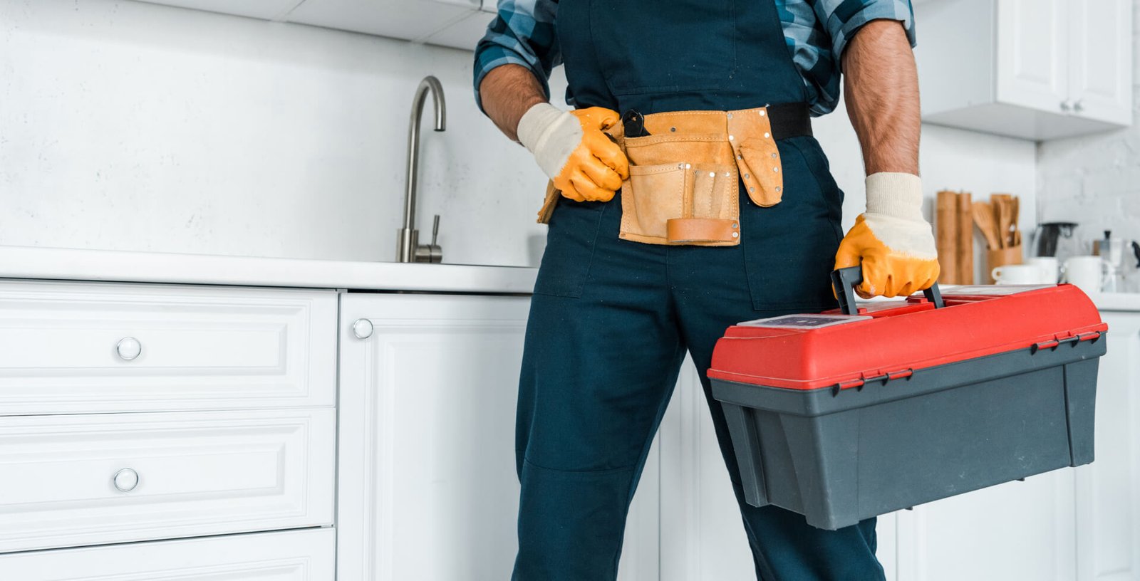 A plumber in work overalls and gloves holds a red toolbox in a modern white kitchen.