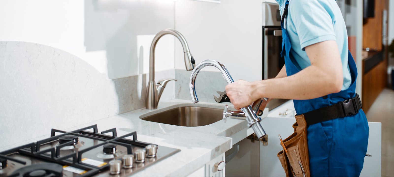 A plumber in blue overalls installs a new chrome faucet in a modern kitchen sink.