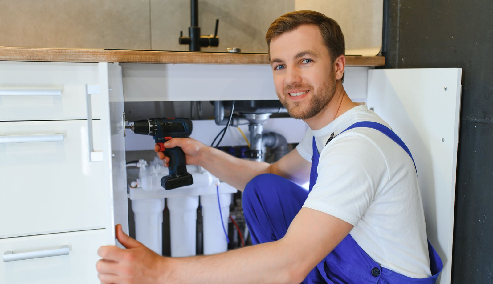 A smiling plumber in blue overalls uses a drill while repairing pipes under a kitchen sink.